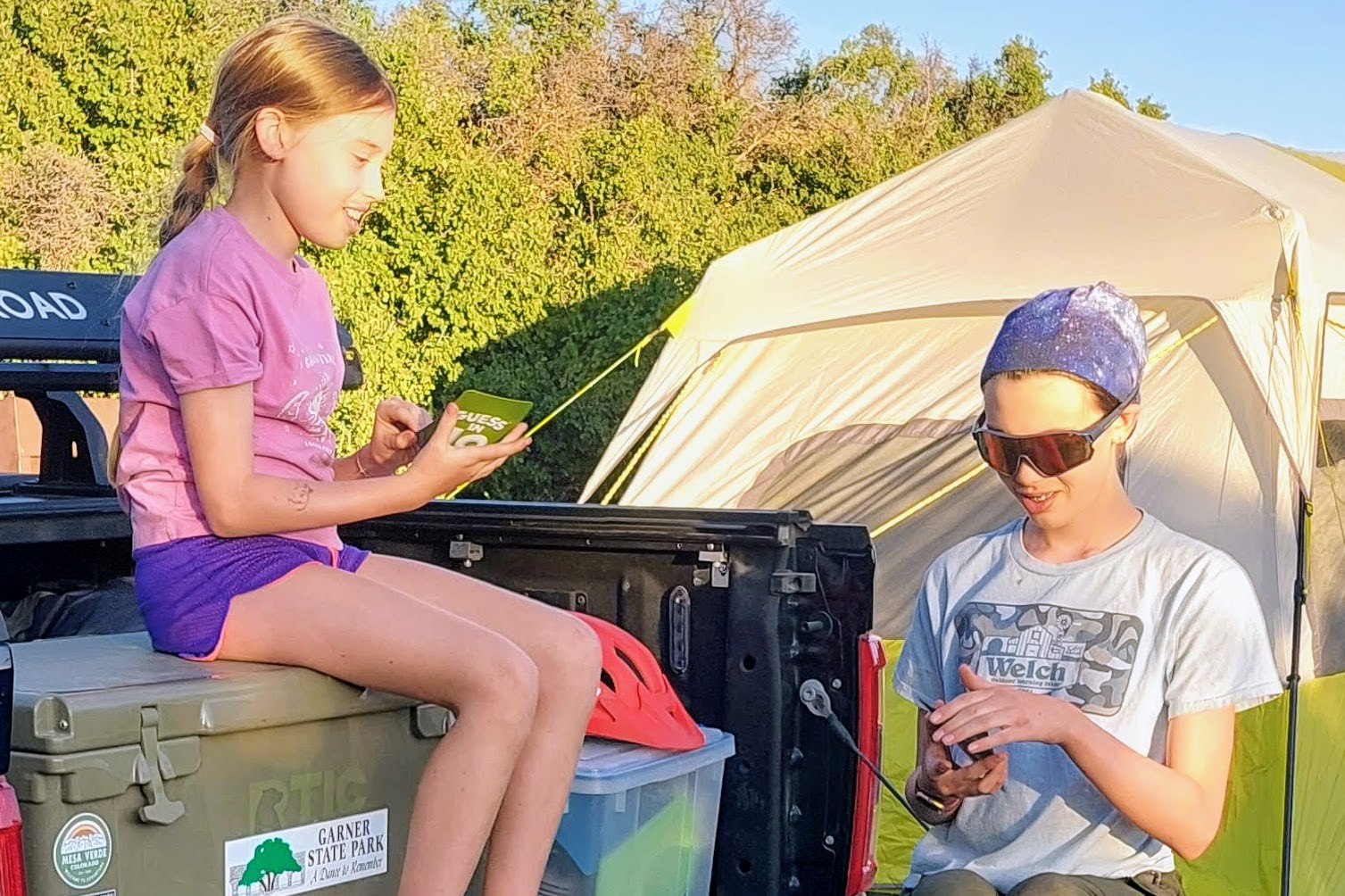 Two children relaxing beside a family camping tent in casual clothes at a campsite during golden hour, showing comfortable clothing for warm weather camping.